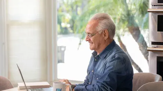An older gentalmen sitting down at a desk, typing on his computer, with a mesawater cup sitting next to his laptop.